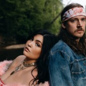 A man and woman stood back to back in front of a river. the woman is wearing a pink fluffy coat around her shoulders and lots of silver necklaces. The man has on a blue denim jacket and pink and white tie dye bandana around his head